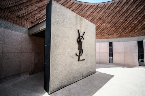 A modern architectural structure with a prominent YSL logo on a textured stone wall. Surrounding the structure is a curved wall made of light stone and brick, adding an architectural contrast. The setting appears to be outdoors, with natural lighting highlighting the materials and design.