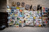 A variety of colorful and artistic signs and masks are displayed against a textured wall. The signs feature illustrations and text in multiple languages, portraying various professions and public services such as police, ambulance, carpenter, and more. Above the signs, decorative tribal masks with bold patterns are arranged.