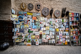 A variety of colorful and artistic signs and masks are displayed against a textured wall. The signs feature illustrations and text in multiple languages, portraying various professions and public services such as police, ambulance, carpenter, and more. Above the signs, decorative tribal masks with bold patterns are arranged.