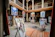 An art gallery with an open central courtyard is filled with various paintings displayed on easels and hanging on walls. The setting includes wooden beams and pillars, giving it a traditional aesthetic. Colorful artwork in different styles is prominently featured.