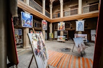 An art gallery with an open central courtyard is filled with various paintings displayed on easels and hanging on walls. The setting includes wooden beams and pillars, giving it a traditional aesthetic. Colorful artwork in different styles is prominently featured.