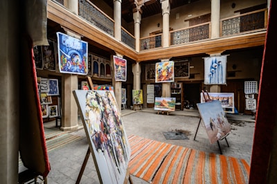 An art gallery with an open central courtyard is filled with various paintings displayed on easels and hanging on walls. The setting includes wooden beams and pillars, giving it a traditional aesthetic. Colorful artwork in different styles is prominently featured.