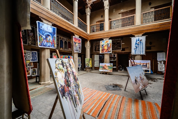 An art gallery with an open central courtyard is filled with various paintings displayed on easels and hanging on walls. The setting includes wooden beams and pillars, giving it a traditional aesthetic. Colorful artwork in different styles is prominently featured.