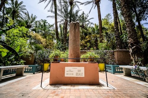 A lush garden area featuring a central raised stone structure with an engraved plaque, surrounded by large potted plants and flanked by large trees. There are benches on either side of the stone structure, creating a serene, memorial-like setting.