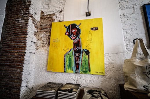 A painting is mounted on a textured white wall, featuring a stylized humanoid figure with a dark body and bright, bold colors against a vivid yellow background. The figure has prominent red eyes and horns, with symbolic and abstract elements surrounding it. Below the painting, a stack of books and a cloth bag are visible, all set against a rustic and partially exposed brick background.
