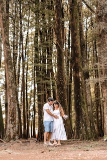 A couple stands in a serene forest setting, surrounded by tall, majestic trees. The woman in a white dress is holding a small bouquet, while the man beside her gently places his hand on her belly, suggesting a pregnancy. The scene exudes warmth and intimacy, with natural light filtering through the trees.