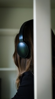 A person wearing large, over-ear headphones is partially obscured by a light-colored vertical object. The headphones are dark, and the person's hair is long and brown. The background is out of focus, displaying muted tones.
