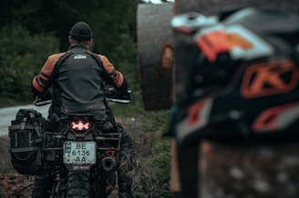 A person wearing a dark gray and orange motorcycle jacket, seated on a motorcycle with a visible license plate and off-road tires. The background is a blurred natural setting with green foliage. Another motorcycle part is visible in the foreground, partially out of focus.