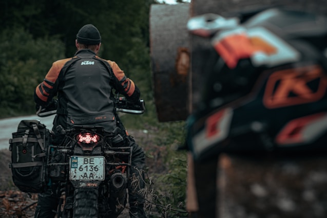 A person wearing a dark gray and orange motorcycle jacket, seated on a motorcycle with a visible license plate and off-road tires. The background is a blurred natural setting with green foliage. Another motorcycle part is visible in the foreground, partially out of focus.