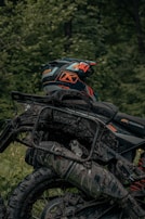 A close-up view of a mud-covered motorcycle with a helmet resting on the seat. The helmet features bright colors and branding, while the bike is parked in a lush, green forest setting. The motorcycle has visible detailing of its suspension and frame, with mud splattered all over the tires and body.