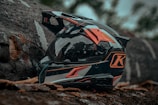 A motorcycle helmet with a sleek design is placed on the ground surrounded by natural elements like tree bark and leaves. The helmet features bold colors and graphics, including prominent orange and black hues.