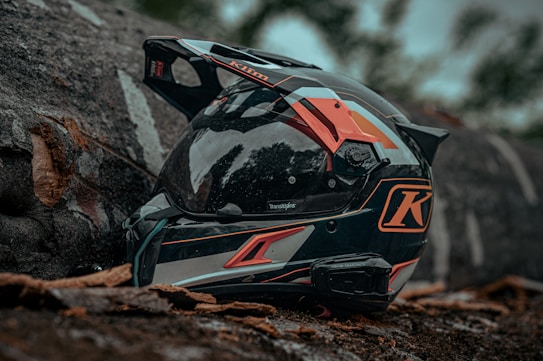 A motorcycle helmet with a sleek design is placed on the ground surrounded by natural elements like tree bark and leaves. The helmet features bold colors and graphics, including prominent orange and black hues.