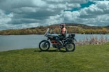 A person in riding gear is seated on a touring motorcycle near a lake, surrounded by lush green grass and trees. The sky is filled with large, dramatic clouds, contributing to a scenic and adventurous setting.