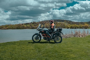 A person in riding gear is seated on a touring motorcycle near a lake, surrounded by lush green grass and trees. The sky is filled with large, dramatic clouds, contributing to a scenic and adventurous setting.