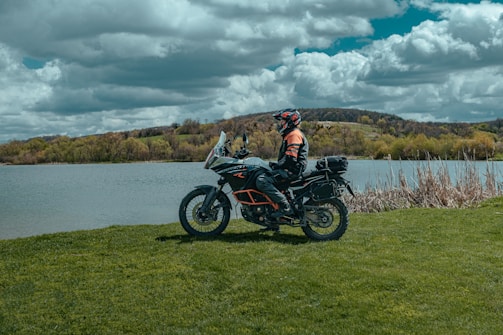 A person in riding gear is seated on a touring motorcycle near a lake, surrounded by lush green grass and trees. The sky is filled with large, dramatic clouds, contributing to a scenic and adventurous setting.