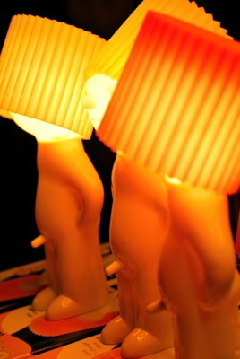 Three uniquely designed lamps shaped like stylized human figures with lampshades resembling cupcake liners are arranged on a surface. The light emitted is warm, creating an orange-yellow glow that casts a soft ambiance.