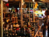 A bustling market stall is packed with a variety of bottled goods, including oils and spirits. A person is browsing the items on display, reaching out towards a bottle. The stall is under a canopy, with colorful tarps and lights enhancing the vibrant atmosphere.