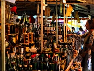 A bustling market stall is packed with a variety of bottled goods, including oils and spirits. A person is browsing the items on display, reaching out towards a bottle. The stall is under a canopy, with colorful tarps and lights enhancing the vibrant atmosphere.