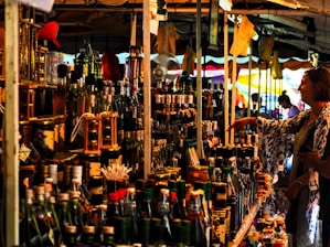 A bustling market stall is packed with a variety of bottled goods, including oils and spirits. A person is browsing the items on display, reaching out towards a bottle. The stall is under a canopy, with colorful tarps and lights enhancing the vibrant atmosphere.