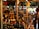A bustling market stall is packed with a variety of bottled goods, including oils and spirits. A person is browsing the items on display, reaching out towards a bottle. The stall is under a canopy, with colorful tarps and lights enhancing the vibrant atmosphere.