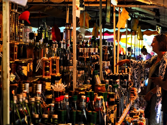 A bustling market stall is packed with a variety of bottled goods, including oils and spirits. A person is browsing the items on display, reaching out towards a bottle. The stall is under a canopy, with colorful tarps and lights enhancing the vibrant atmosphere.