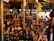 A bustling market stall is packed with a variety of bottled goods, including oils and spirits. A person is browsing the items on display, reaching out towards a bottle. The stall is under a canopy, with colorful tarps and lights enhancing the vibrant atmosphere.