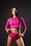 A woman stands confidently against a dark background wearing a bright pink long-sleeve crop top and matching shorts. Her hair is long and loose, and her lips are painted with a bold color, adding to her assertive expression. The overall pose and attire suggest a sense of empowerment and strength.