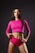 A woman stands confidently against a dark background wearing a bright pink long-sleeve crop top and matching shorts. Her hair is long and loose, and her lips are painted with a bold color, adding to her assertive expression. The overall pose and attire suggest a sense of empowerment and strength.