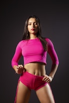 A woman stands confidently against a dark background wearing a bright pink long-sleeve crop top and matching shorts. Her hair is long and loose, and her lips are painted with a bold color, adding to her assertive expression. The overall pose and attire suggest a sense of empowerment and strength.