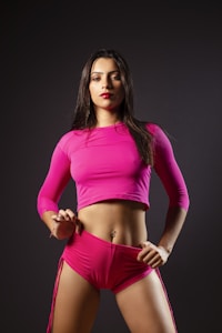 A woman stands confidently against a dark background wearing a bright pink long-sleeve crop top and matching shorts. Her hair is long and loose, and her lips are painted with a bold color, adding to her assertive expression. The overall pose and attire suggest a sense of empowerment and strength.