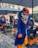 Participants in vibrant carnival costumes are riding uniquely designed bikes in a street parade. The costumes feature large, exaggerated masks with bold expressions and vivid blue and orange colors. Confetti covers the ground, and large umbrellas provide shade overhead.