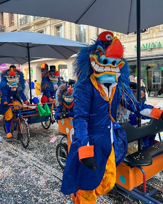 Participants in vibrant carnival costumes are riding uniquely designed bikes in a street parade. The costumes feature large, exaggerated masks with bold expressions and vivid blue and orange colors. Confetti covers the ground, and large umbrellas provide shade overhead.