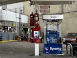 A gas station with a Mobil fuel pump displaying regular, extra, and supreme options. Two red fire extinguishers are mounted above the pump. The area is surrounded by buildings and contains a red vehicle parked nearby. An ATM machine is visible with several signs, including warnings and price listings.