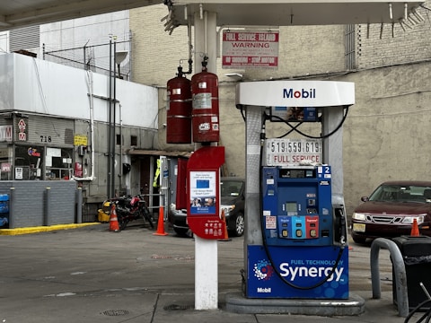 A gas station with a Mobil fuel pump displaying regular, extra, and supreme options. Two red fire extinguishers are mounted above the pump. The area is surrounded by buildings and contains a red vehicle parked nearby. An ATM machine is visible with several signs, including warnings and price listings.