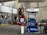 A gas station with a Mobil fuel pump displaying regular, extra, and supreme options. Two red fire extinguishers are mounted above the pump. The area is surrounded by buildings and contains a red vehicle parked nearby. An ATM machine is visible with several signs, including warnings and price listings.