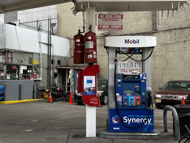 A gas station with a Mobil fuel pump displaying regular, extra, and supreme options. Two red fire extinguishers are mounted above the pump. The area is surrounded by buildings and contains a red vehicle parked nearby. An ATM machine is visible with several signs, including warnings and price listings.