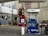 A gas station with a Mobil fuel pump displaying regular, extra, and supreme options. Two red fire extinguishers are mounted above the pump. The area is surrounded by buildings and contains a red vehicle parked nearby. An ATM machine is visible with several signs, including warnings and price listings.