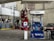 A gas station with a Mobil fuel pump displaying regular, extra, and supreme options. Two red fire extinguishers are mounted above the pump. The area is surrounded by buildings and contains a red vehicle parked nearby. An ATM machine is visible with several signs, including warnings and price listings.