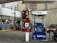 A gas station with a Mobil fuel pump displaying regular, extra, and supreme options. Two red fire extinguishers are mounted above the pump. The area is surrounded by buildings and contains a red vehicle parked nearby. An ATM machine is visible with several signs, including warnings and price listings.