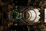 An intricately designed indoor space features an ornate glass ceiling with arched frameworks and decorative detailing. The interior includes abundant greenery, surrounded by comfortable seating arrangements. The use of warm lighting highlights the elaborate aesthetics and adds to the luxurious ambiance.
