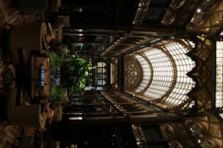 An intricately designed indoor space features an ornate glass ceiling with arched frameworks and decorative detailing. The interior includes abundant greenery, surrounded by comfortable seating arrangements. The use of warm lighting highlights the elaborate aesthetics and adds to the luxurious ambiance.