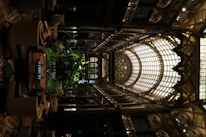 An intricately designed indoor space features an ornate glass ceiling with arched frameworks and decorative detailing. The interior includes abundant greenery, surrounded by comfortable seating arrangements. The use of warm lighting highlights the elaborate aesthetics and adds to the luxurious ambiance.