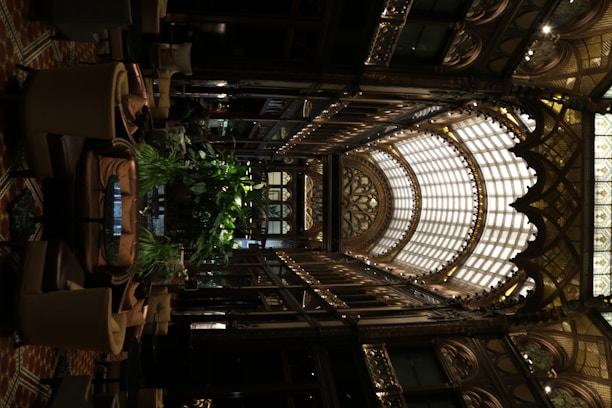 An intricately designed indoor space features an ornate glass ceiling with arched frameworks and decorative detailing. The interior includes abundant greenery, surrounded by comfortable seating arrangements. The use of warm lighting highlights the elaborate aesthetics and adds to the luxurious ambiance.