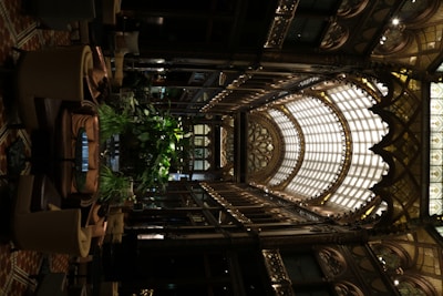 An intricately designed indoor space features an ornate glass ceiling with arched frameworks and decorative detailing. The interior includes abundant greenery, surrounded by comfortable seating arrangements. The use of warm lighting highlights the elaborate aesthetics and adds to the luxurious ambiance.