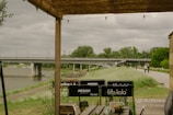 A wooden pergola provides partial shelter to an outdoor seating area with various branded signs on the chairs. In the background, there's a wide river crossed by a large bridge. The overcast sky adds a moody atmosphere, while trees and a grassy path line the riverbank.