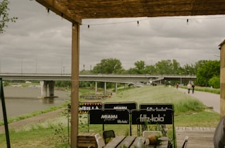 A wooden pergola provides partial shelter to an outdoor seating area with various branded signs on the chairs. In the background, there's a wide river crossed by a large bridge. The overcast sky adds a moody atmosphere, while trees and a grassy path line the riverbank.