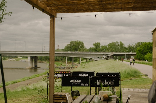 A wooden pergola provides partial shelter to an outdoor seating area with various branded signs on the chairs. In the background, there's a wide river crossed by a large bridge. The overcast sky adds a moody atmosphere, while trees and a grassy path line the riverbank.