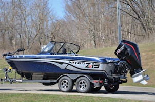 A blue and black Nitro Z19 sport fishing boat is mounted on a trailer. The boat features a sleek, modern design with a powerful Mercury Pro XS outboard motor at the rear. The setting appears to be a wooded area with leafless trees, likely indicating early spring or late fall.