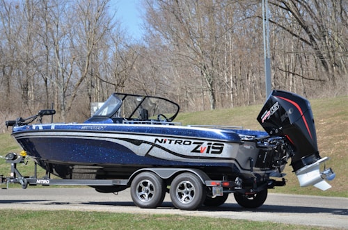 A blue and black Nitro Z19 sport fishing boat is mounted on a trailer. The boat features a sleek, modern design with a powerful Mercury Pro XS outboard motor at the rear. The setting appears to be a wooded area with leafless trees, likely indicating early spring or late fall.