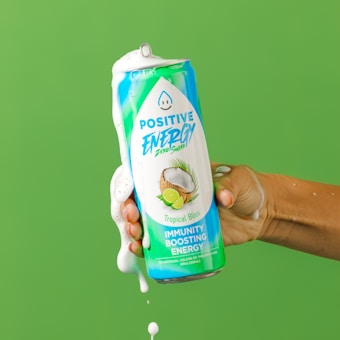 A hand holding a can of energy drink with liquid overflowing and foaming from the top. The can is labeled 'Positive Energy' with a tropical theme, displaying images of a coconut and lime.